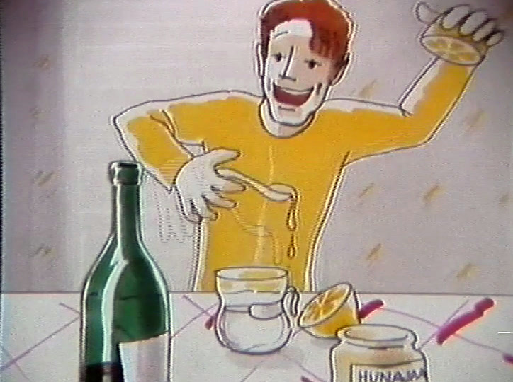 At a table with a green bottle and a jar marked HUNAJA, he holds up a lemon.
