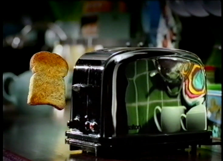 Toast leaps out of a black toaster on a kitchen counter, teapot blurred behind