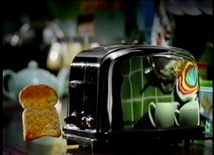 Two slices hang in the air beside the toaster, the green kitchen bent across its chrome flank
