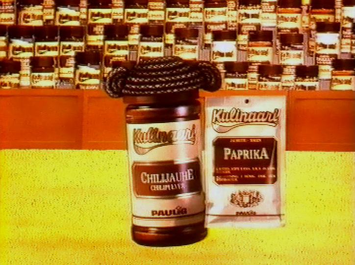 A chilli powder jar and a paprika sachet in front of a wall stacked with spice jars.