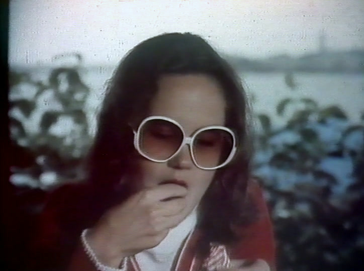 A woman in a red coat unwraps a sweet by the water, large round sunglasses on.