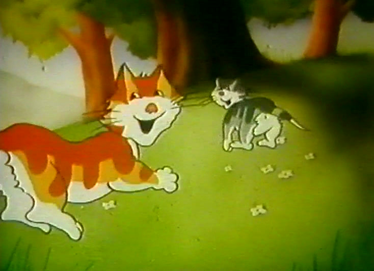 A cat sits on a picnic rug in a meadow, basket and filled glasses beside it.