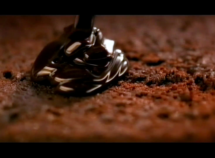 A spatula drags dark chocolate icing across a cake dusted with crumbs