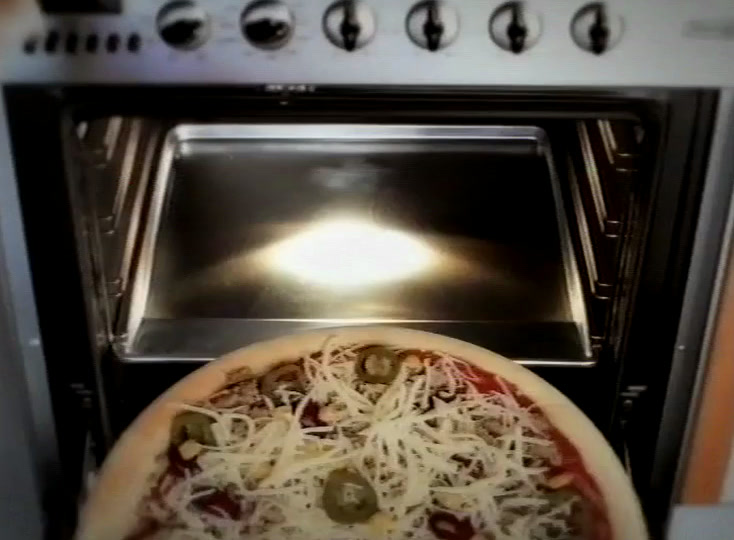 A topped pizza slides into a lit domestic oven, dials in a row above the door