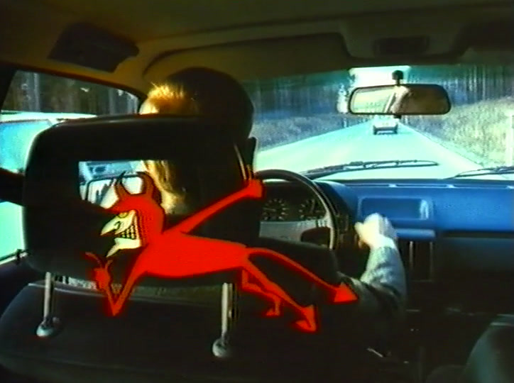 Crouched low, the devil points through the windscreen at a pale saloon car on the road.