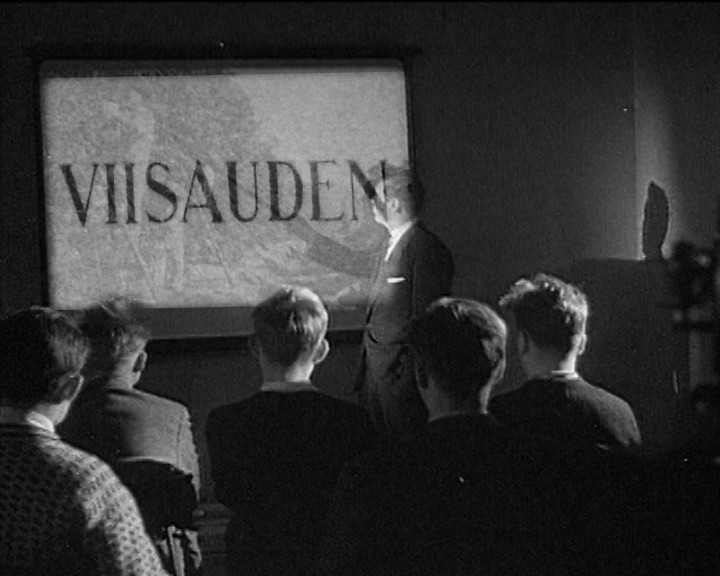 Boys watch a man beside a projection screen in a dark room; the screen reads "VIISAUDEN".