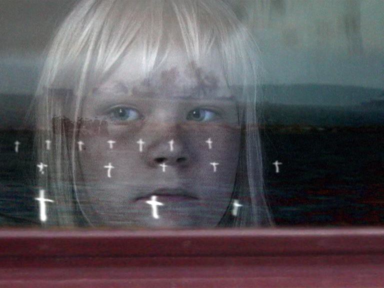 A child's face behind glass, rows of small white crosses reflected across it.