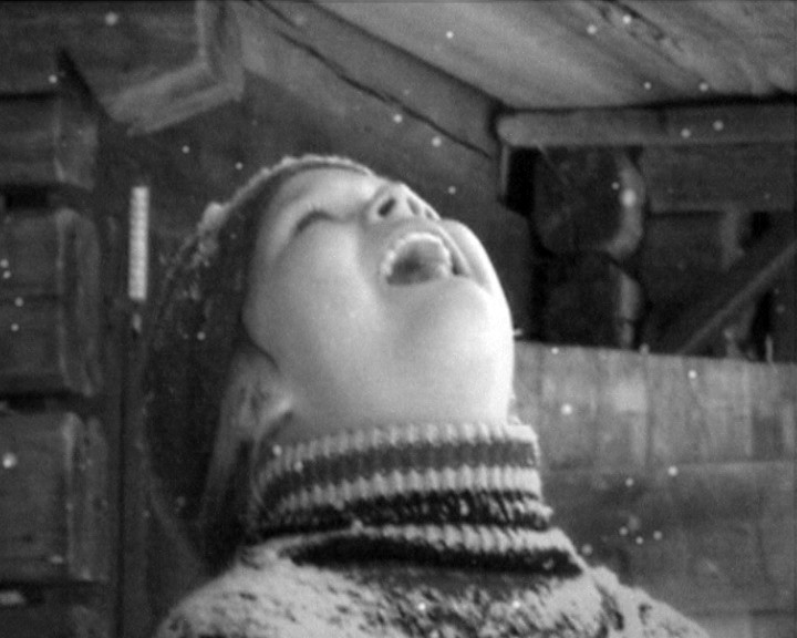 Close-up of a child in a knitted hat, head tipped back, tongue out to catch snow.