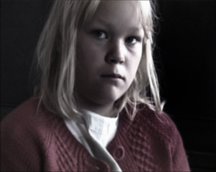 A fair-haired child in a red knitted cardigan and white collar looks past the camera.