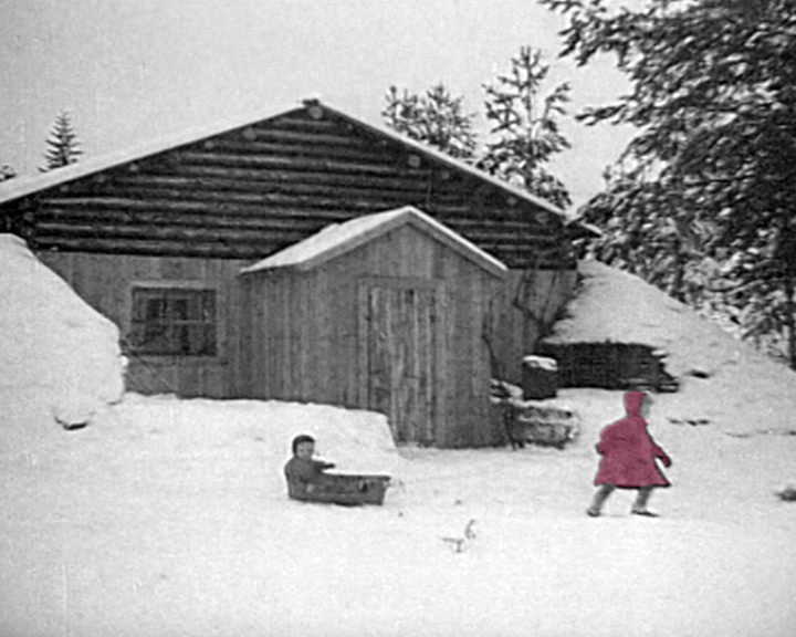 A child sits in a wooden sledge in the snow outside a log house, another walks past in red.
