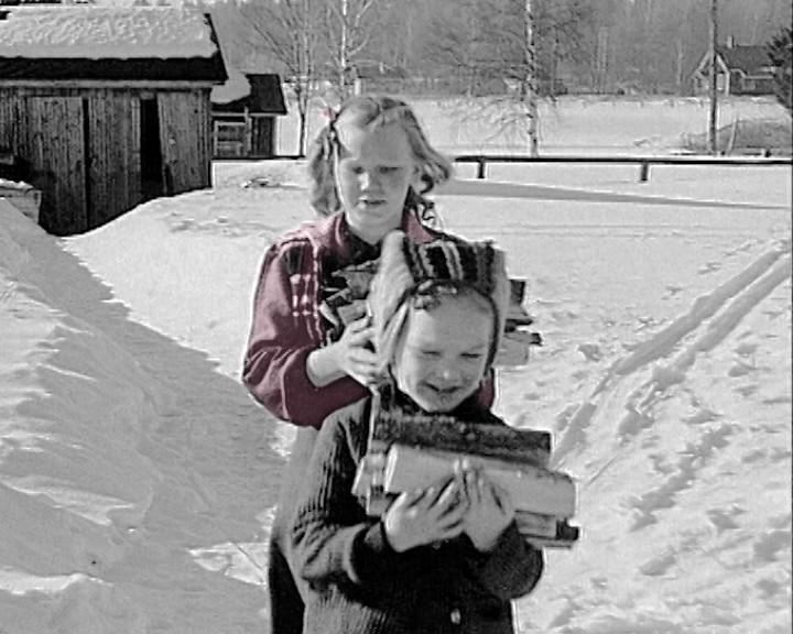 Two children carry armfuls of firewood across deep snow past a wooden barn.