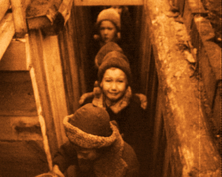 Children in knitted hats climb one behind another out of a narrow passage between rough timber walls.