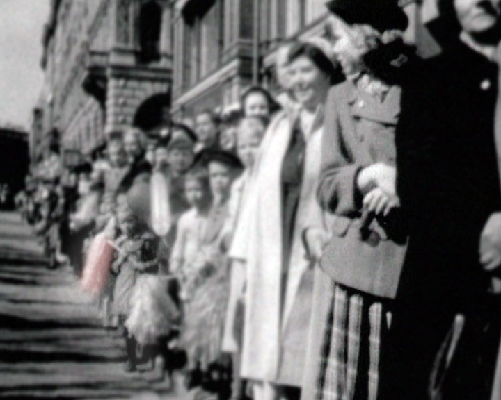 A crowd of women and children in coats and hats lines a street outside a stone building.