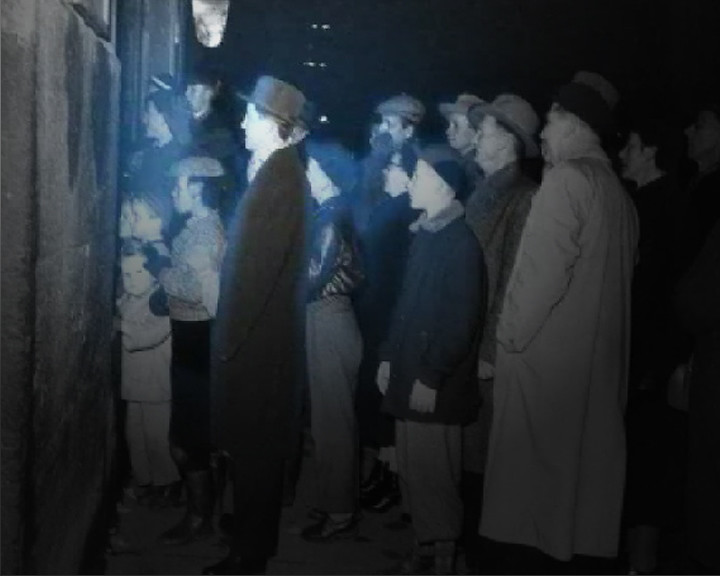A crowd of adults and children in coats and hats queues along a wall, tinted blue.