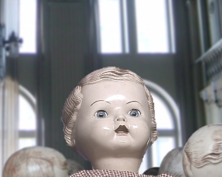An old doll with moulded hair and blue eyes stands before tall windows, other dolls behind.