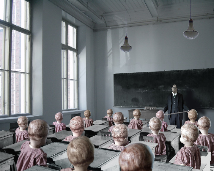 Rows of dolls in pink dresses fill a classroom; a man with a pointer stands at the blackboard.