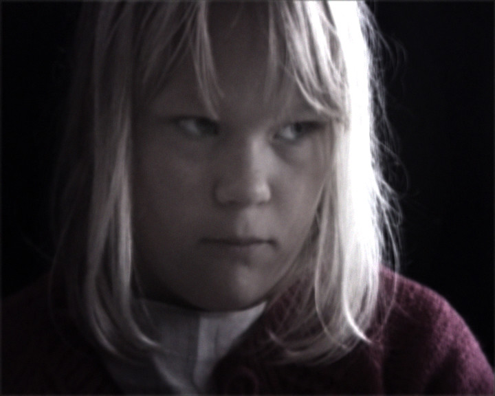 A fair-haired child in a red cardigan looks sideways, lit against a black background.