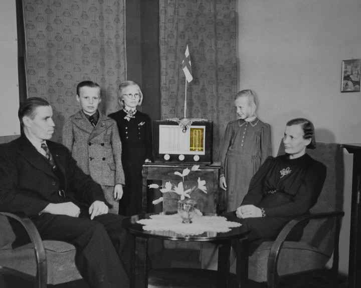 A family of five poses in a living room around a radio topped with a small Finnish flag.