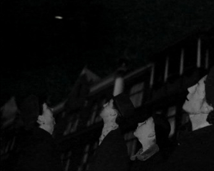 Several people in hats and dark coats stand in near darkness, their lit faces in profile.