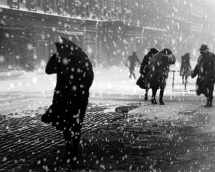 People in coats and hats push through heavy snowfall on a city street, one carrying an umbrella.