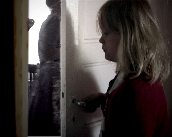 A girl in a red cardigan holds a door handle while a blurred figure stands beyond.