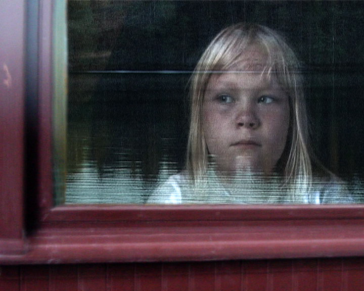 A fair-haired girl looks out through a red-framed window, water reflected in the glass.