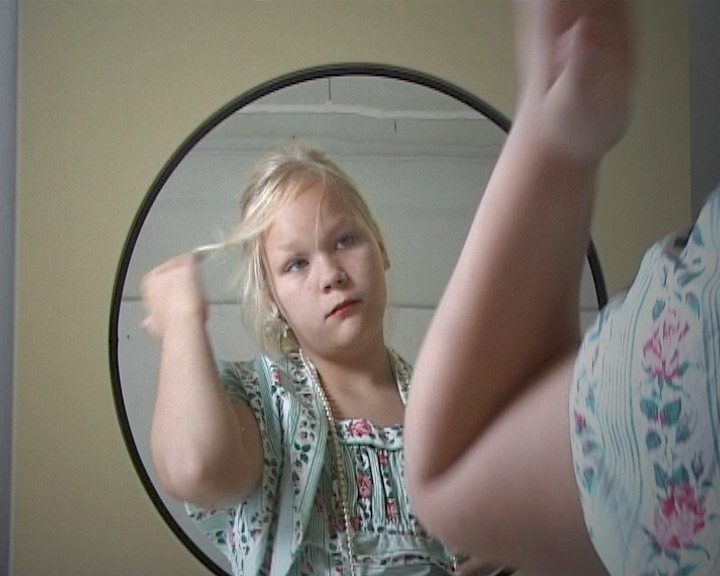 A girl in a pearl necklace raises a hand to her hair in front of a round mirror.