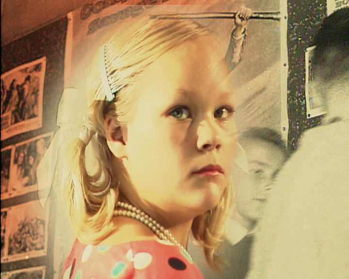 A girl in pearls and a polka-dot dress looks aside; pinned photographs cover the wall behind her.