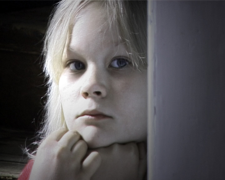 A fair-haired girl rests her chin on her hands beside a pale doorframe, looking out.