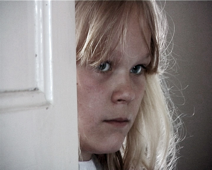 A fair-haired girl looks straight at the camera from behind the edge of a white door frame.