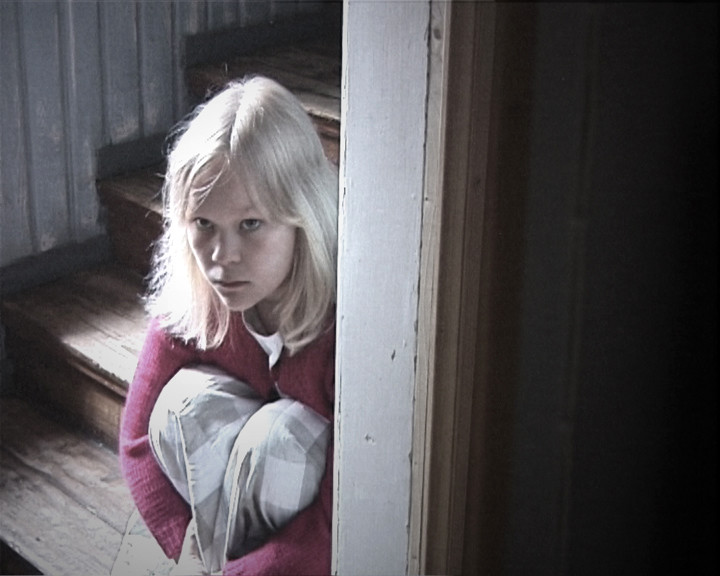 A fair-haired girl in a red cardigan hugs a pillow on wooden stairs, half behind a door frame.