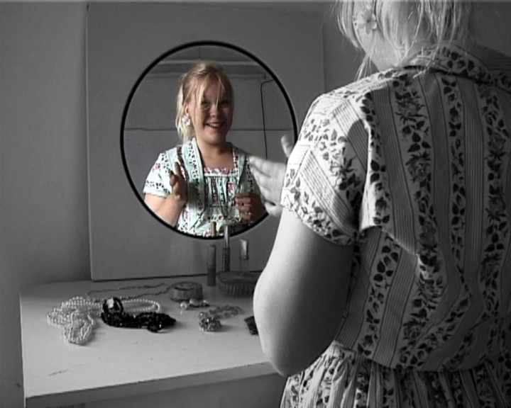 A round mirror reflects a smiling girl in a patterned dress, the only colour in a grey room.