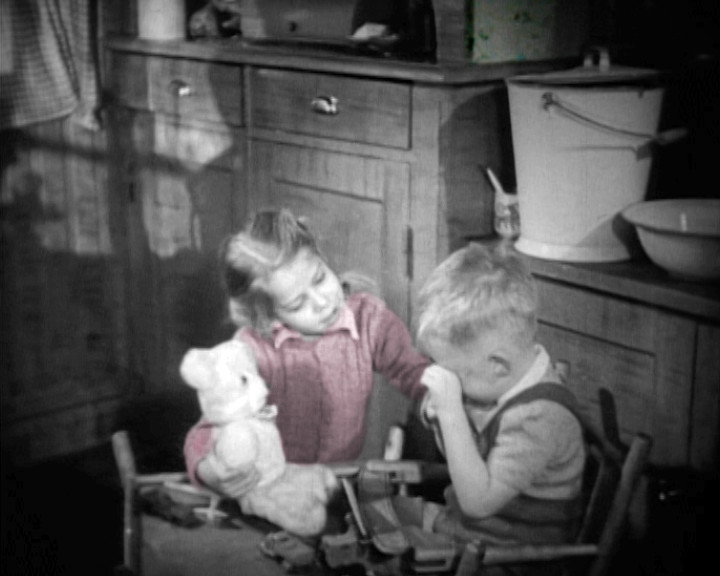 A girl in a red jumper holds a white teddy bear beside a boy in a kitchen.