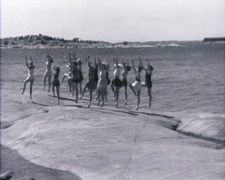 Girls in swimsuits jump with their arms raised on a flat rock beside the sea.