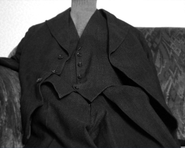 A dark suit jacket and waistcoat with no head above the collar, on a patterned sofa.