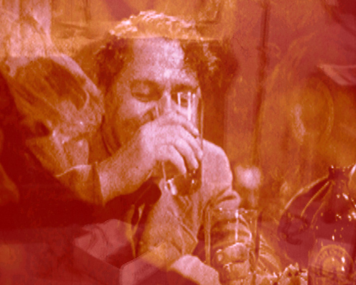 Tinted red, a man with tousled hair drinks from a glass among bottles on a table.