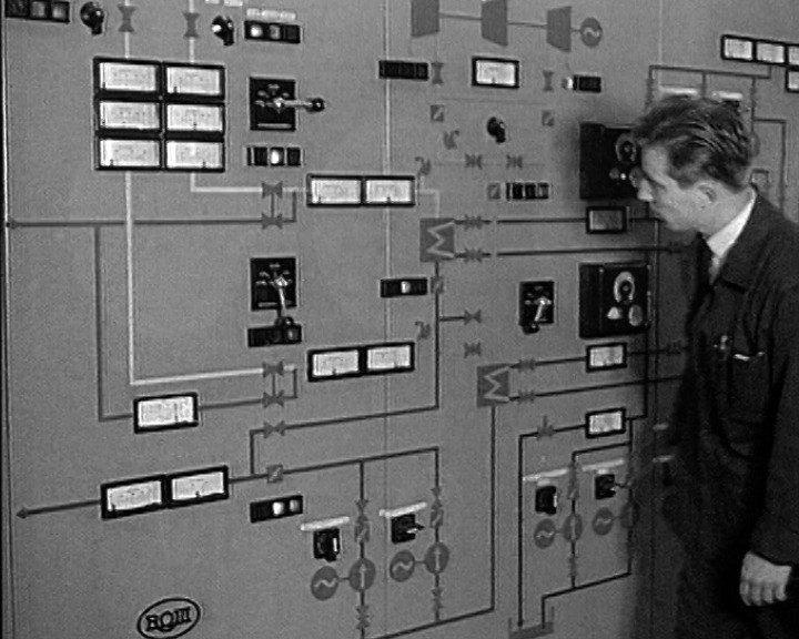 A man in work overalls stands beside a large control panel covered with dials and painted pipework diagrams.