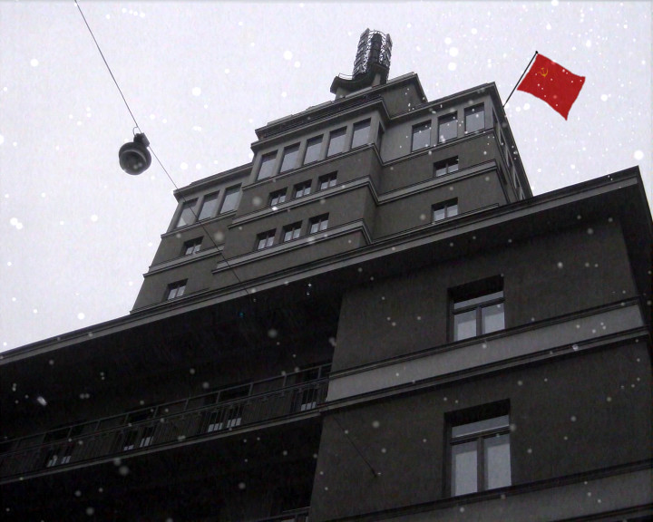 Snow falls past a tall stepped building; a red flag with hammer and sickle flies from the roof.