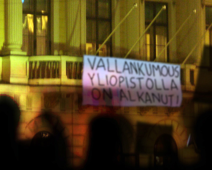 A banner on a floodlit columned building reads “Vallankumous yliopistolla on alkanut!” above silhouetted heads.