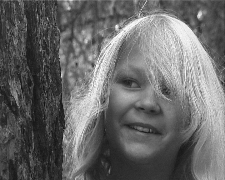 A young girl with long pale hair smiles, half hidden beside the bark of a tree.