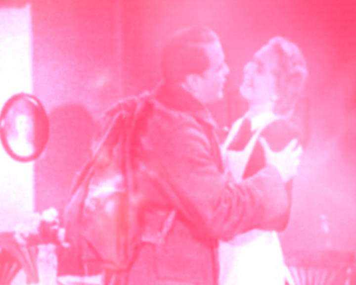 A soldier with a rucksack embraces a woman in a room, the whole frame tinted pink.
