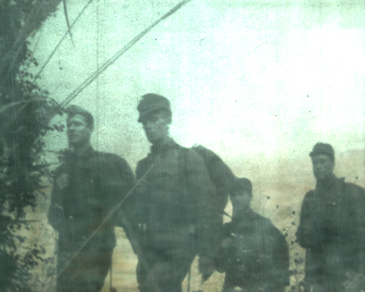 Four soldiers in field caps and long coats stand among bare branches in pale misty light.