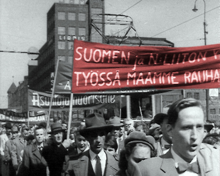 Marchers fill a city street beneath a red banner with white Finnish lettering, partly cut off.