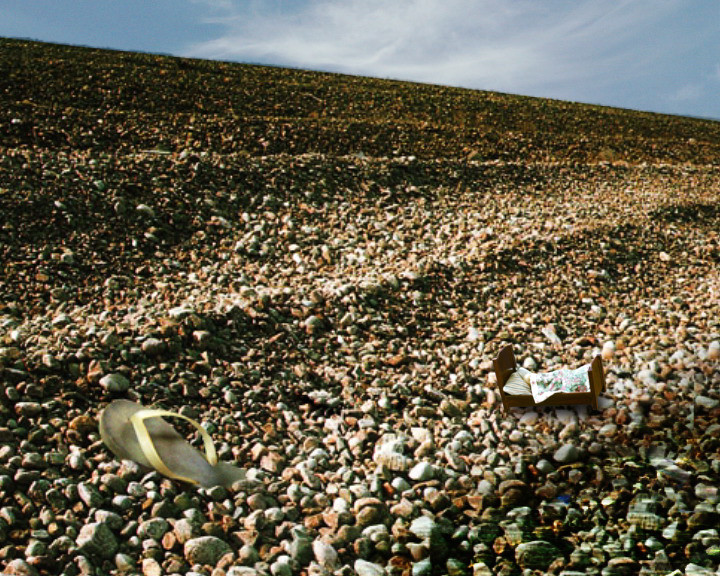 A small wooden bed with a floral quilt and a flip-flop lie on a shingle beach.