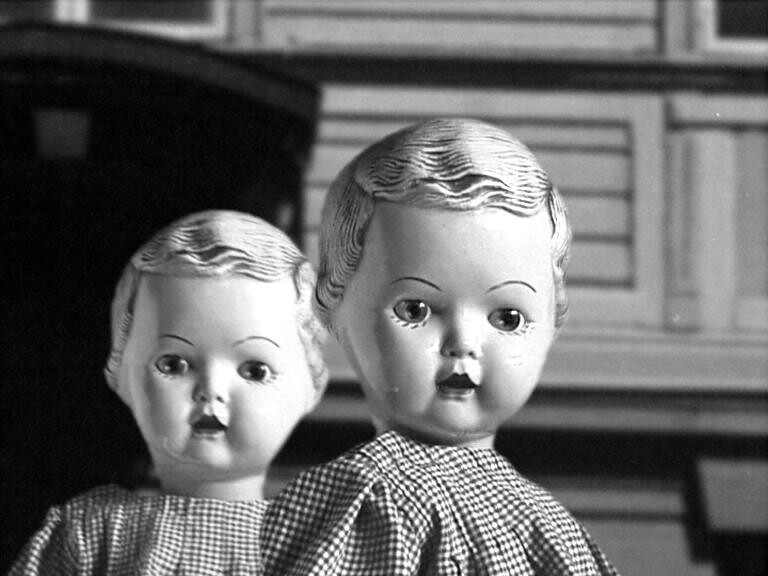 Two dolls in checked dresses stand close together, wood panelling behind them, in black and white.