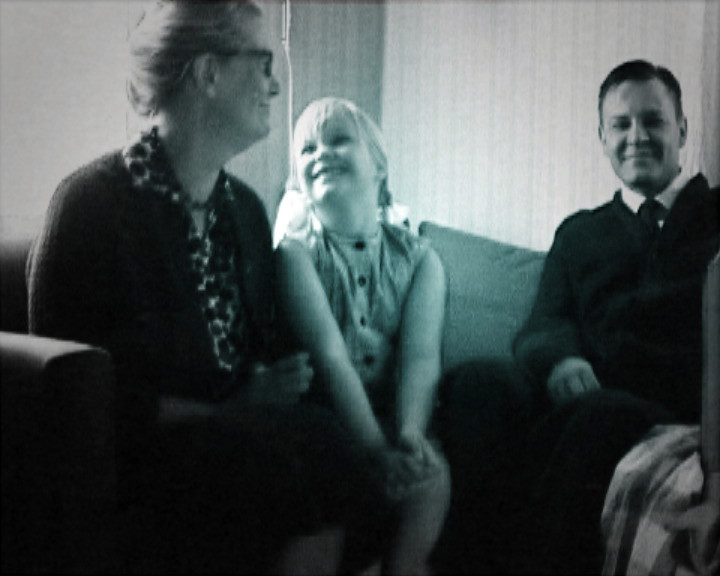 A woman, a laughing young girl and a man in a suit sit together on a sofa.