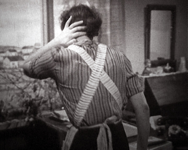 Seen from behind, a woman in a striped blouse and crossed apron straps lifts both hands to her hair.