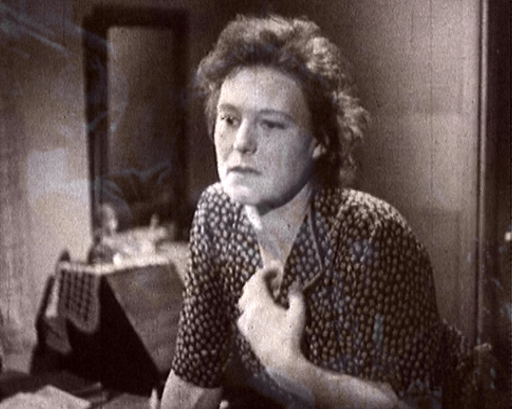 A young woman in a patterned dress sits with one hand at her collar, looking away.