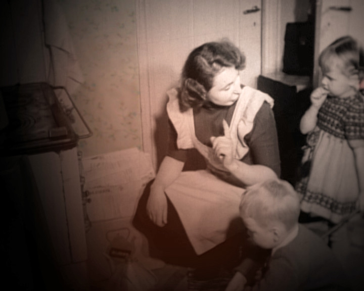 A woman in an apron sits in a corner and raises one finger, two children beside her.