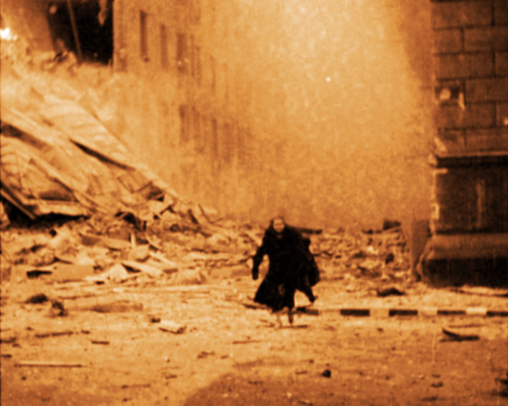 A woman in a dark coat walks towards the camera down a street strewn with rubble beside bombed buildings.